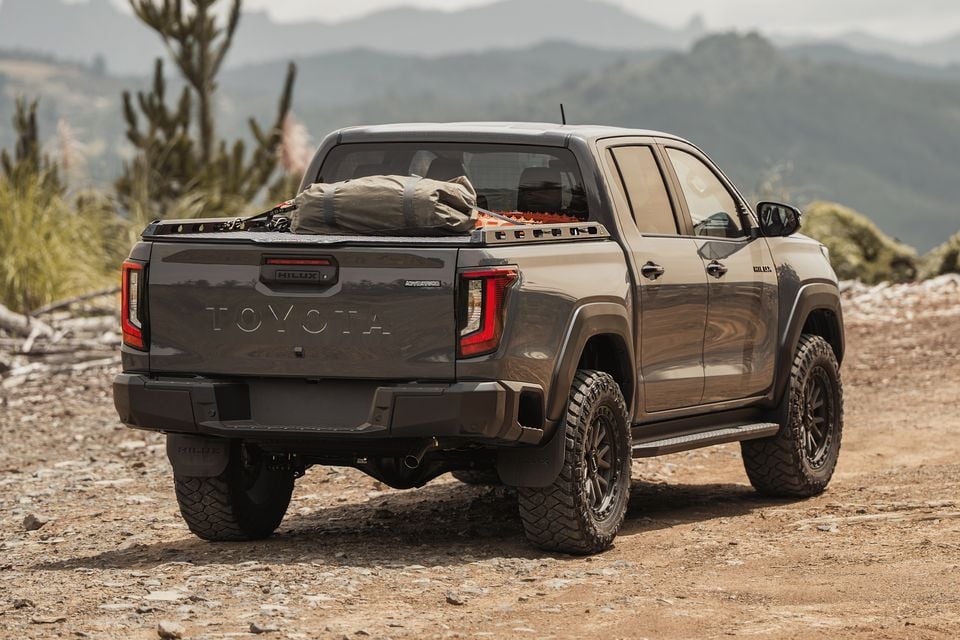 Rear three-quarter view of a grey Toyota HiLux Adventure ute loaded with camping gear, parked on a rocky dirt track with mountains in the background