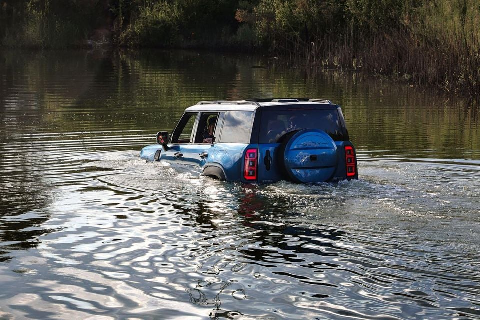 A blue Geely Galaxy Cruiser SUV wading through deep water, seen from the rear three-quarter angle, with a spare wheel mounted on the tailgate