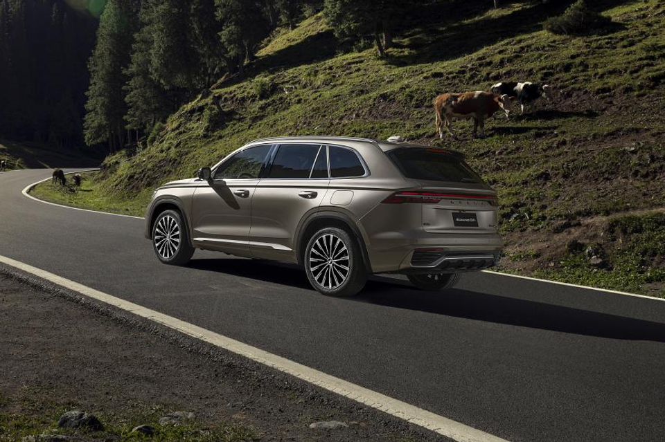 A bronze Geely Monjaro SUV seen from the rear three-quarter angle, driving along a winding mountain road with cows grazing on a hillside in the background.
