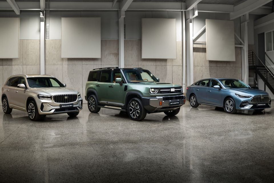Three Geely vehicles displayed indoors: the Monjaro EM-i SUV in silver on the left, the Battleship 700 off-road SUV in green in the centre, and the Emgrand L EM-i sedan in blue on the right.