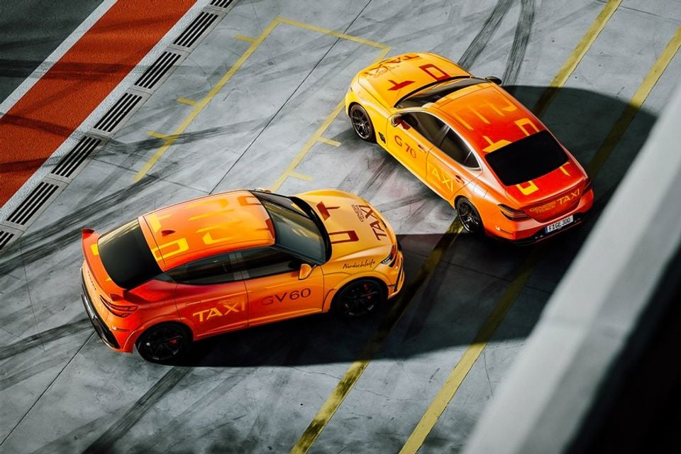 Overhead view of two Genesis Nurburgring Nordschleife taxi cars — a GV60 SUV and a G70 sedan — in bold orange and yellow livery on a race track pit lane