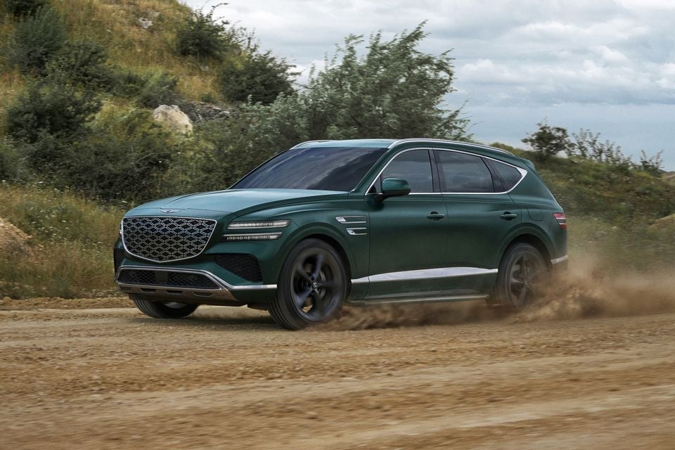 A dark green Genesis GV80 SUV driving on a dirt road, seen from the front-side angle with dust kicking up behind it