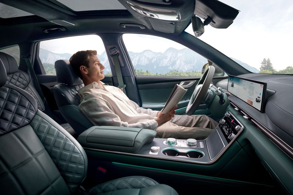 A man reclines in the driver's seat of a Genesis GV80 hybrid, reading a magazine with the seat in a relaxed position, surrounded by quilted leather upholstery and a large touchscreen infotainment display