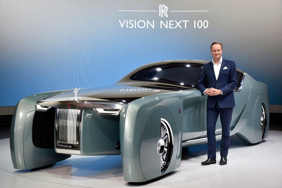 A man in a suit stands beside the Rolls-Royce Vision Next 100 concept car, a sleek grey two-seater shown at an unveiling event