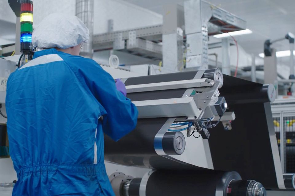 A worker in a blue cleanroom suit and hairnet operating a battery electrode coating machine at a GM battery prototyping facility