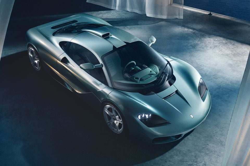 A silver-green Gordon Murray S1 supercar coupe photographed from a high front-three-quarter angle in a moody studio setting, with the dihedral door open revealing a centre-seat interior
