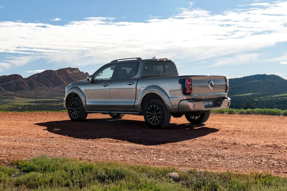 A silver GWM Cannon Alpha dual-cab ute seen from the rear three-quarter angle, parked on a red dirt clearing with mountains in the background.