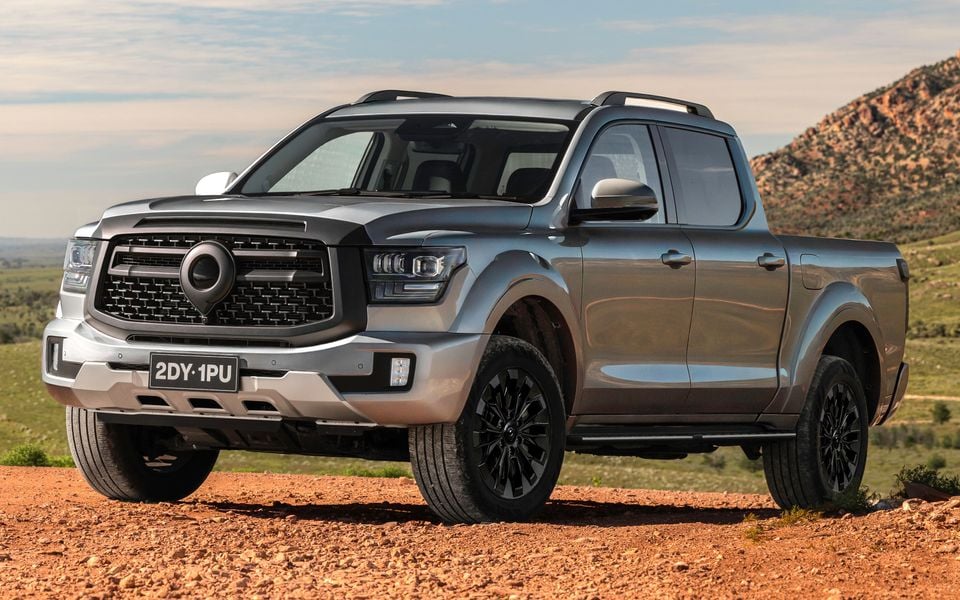 A grey GWM Cannon Alpha dual-cab ute seen from the front-side angle, parked on a red dirt track with rocky hills in the background