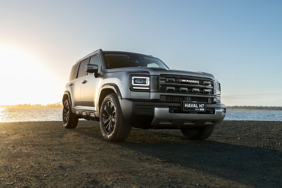 A silver Haval H7 Hi4 PHEV SUV seen from the front-left, parked on a dark gravel surface near a waterway at sunset