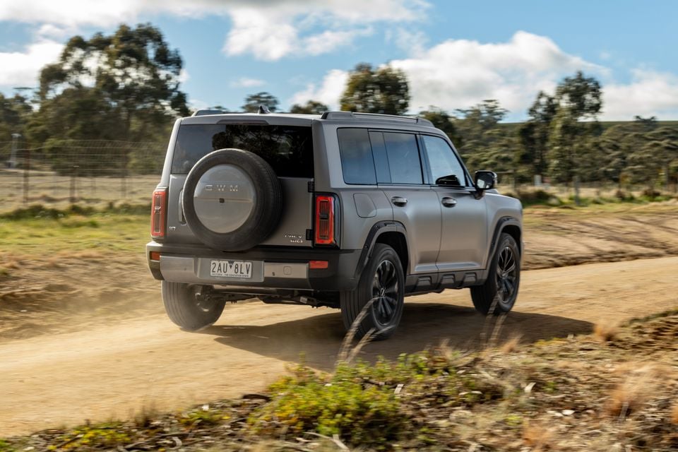 A grey GWM Haval H7 SUV seen from the rear three-quarter angle, driving along a dusty dirt road in a rural Australian setting with a rear-mounted spare wheel visible