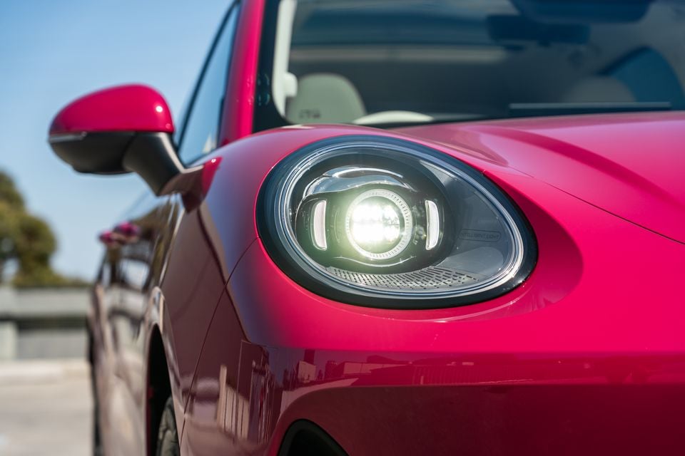 Close-up of the illuminated round LED headlight on a pink GWM Ora 5, with the side mirror and windscreen behind it