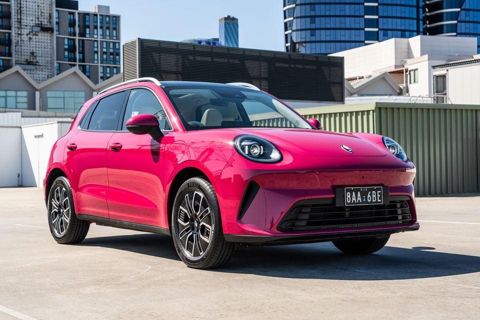 A pink GWM Ora 5 small electric SUV parked on a rooftop car park, seen from the front passenger side with city buildings behind