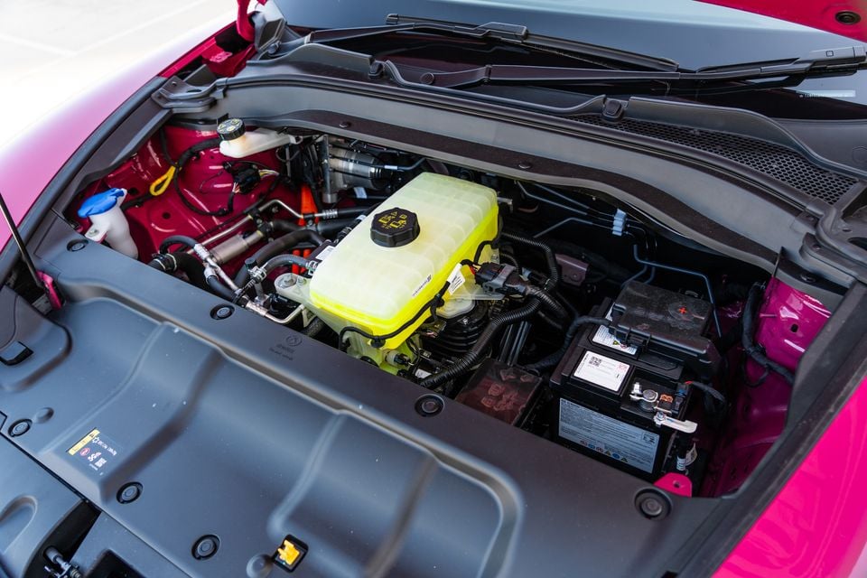 Under the bonnet of a pink GWM Ora 5, showing the coolant reservoir, 12-volt battery and wiring beneath a plastic cover