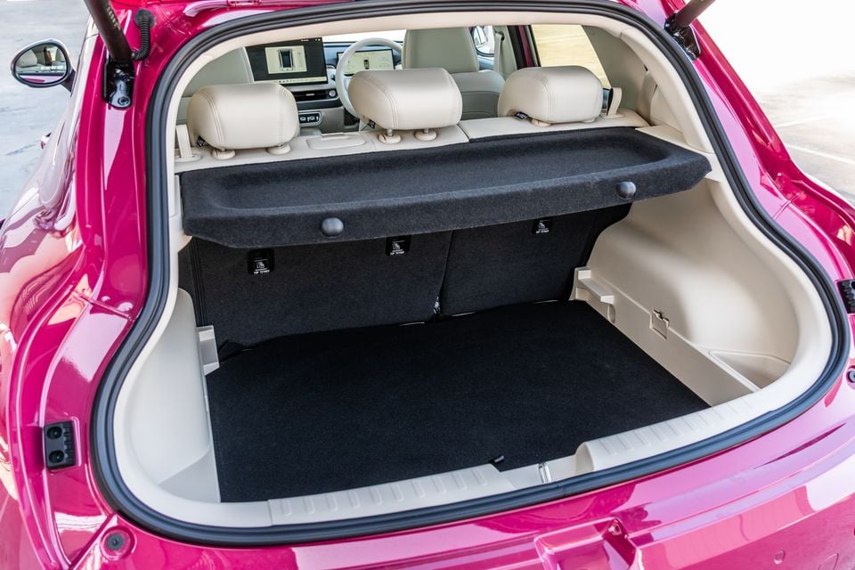 Open boot of a pink GWM Ora 5 showing the carpeted cargo floor, parcel shelf and cream rear seats, with the dashboard screens beyond