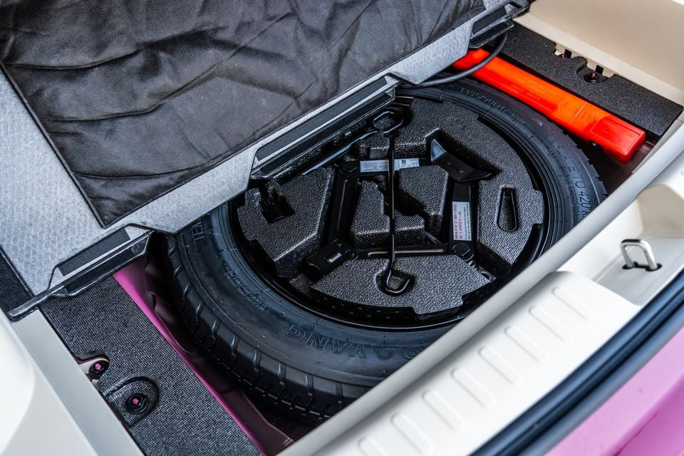 Space-saver spare wheel and jack stored under the boot floor of a GWM Ora 5, with a red warning triangle beside it and pink paintwork visible.