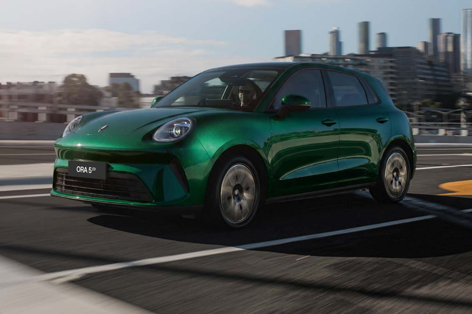 A green GWM ORA 5 EV hatchback driving on a road with a city skyline in the background, seen from the front-side