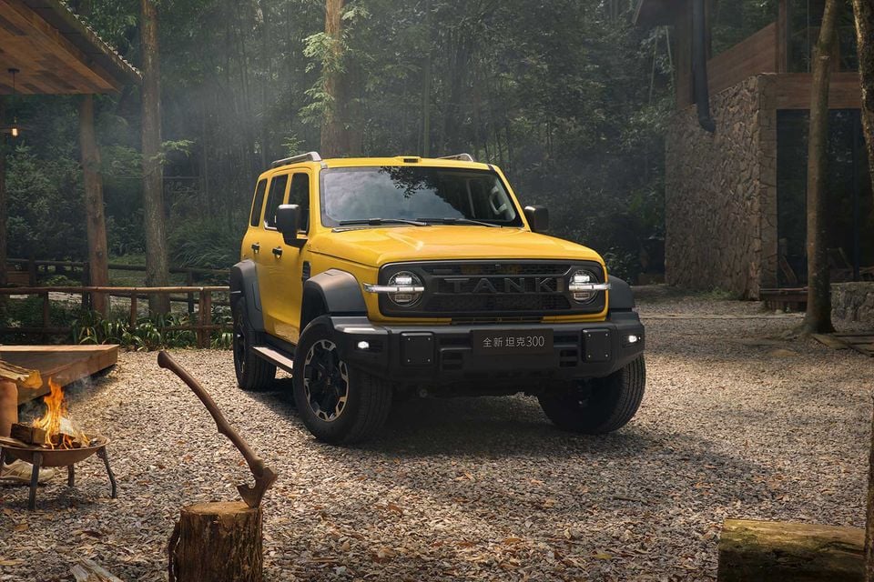 A yellow GWM Tank 300 4WD SUV seen from the front, parked on gravel in a forest setting with a campfire nearby.