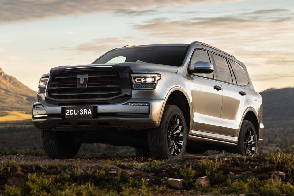 A silver GWM Tank 500 large SUV seen from the front-side angle, parked on rocky high ground with mountains and a sunset sky in the background