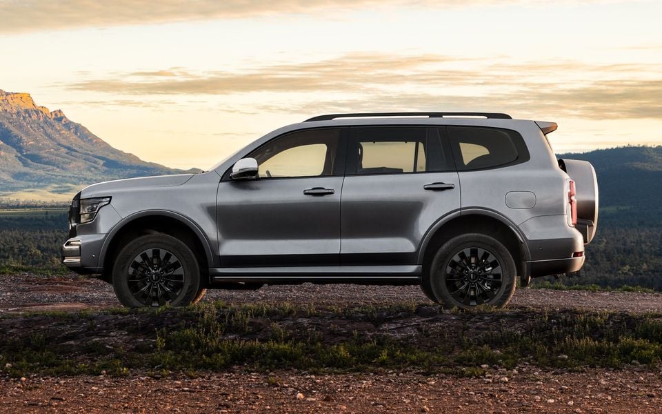 A silver GWM Tank 500 large SUV seen from the side, parked on elevated rocky terrain with mountains and a sunset sky in the background.