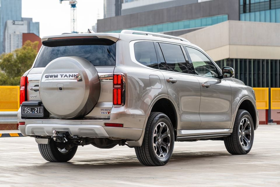 A GWM Tank 500 PHEV SUV seen from the rear three-quarter angle in a sandy/champagne colour, showing the spare wheel, tail lights and tow bar, with a city backdrop