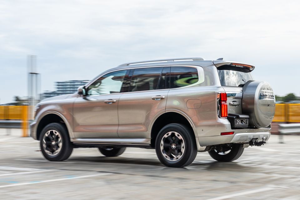 A GWM Tank 500 large SUV seen from the rear three-quarter angle in a car park, finished in a champagne/bronze colour with a rear-mounted spare wheel