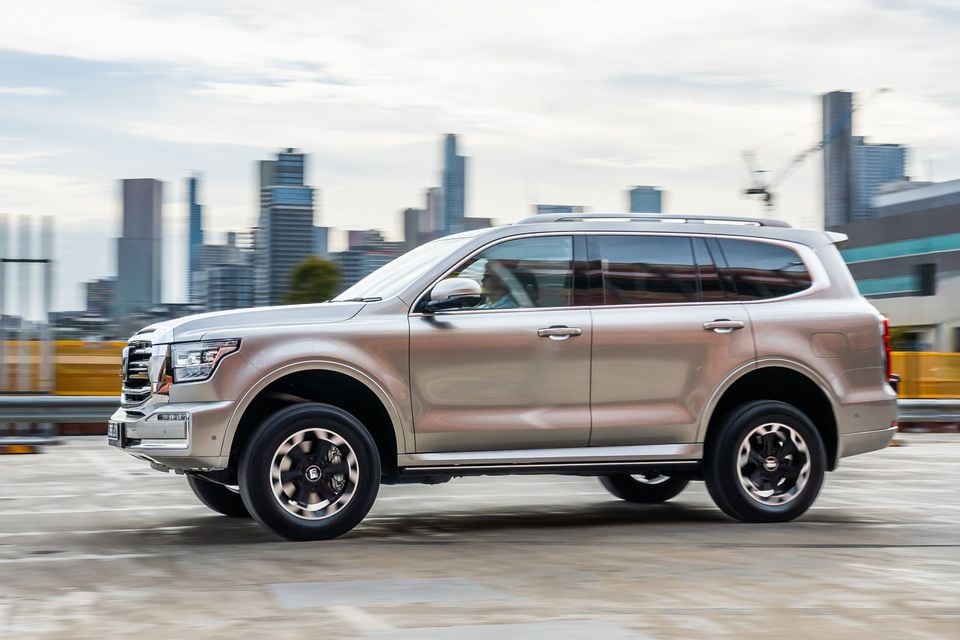 A gold/champagne GWM Tank 500 large SUV seen from the side while driving through a city environment with a skyline in the background