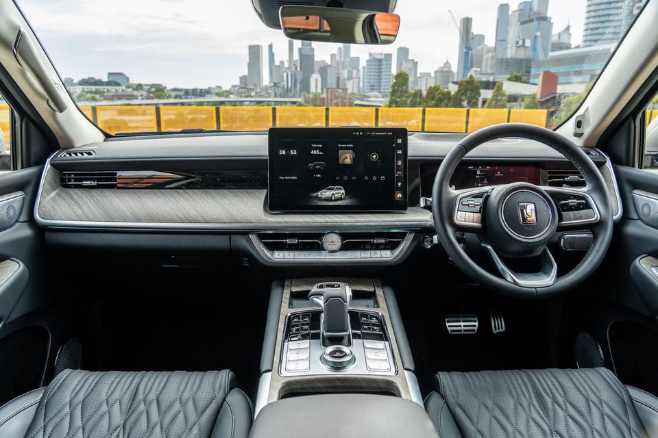 Interior view of a GWM Tank 500 Hi4-T PHEV showing the dashboard, large central touchscreen displaying range and vehicle info, steering wheel, centre console, and quilted leather seats, with the Melbourne city skyline visible through the windscreen