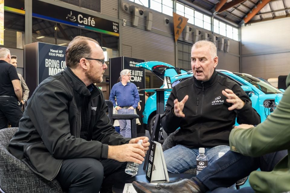 Two men in conversation at a car show, seated in front of a teal GWM Ora electric hatchback with its bonnet open at an indoor exhibition.