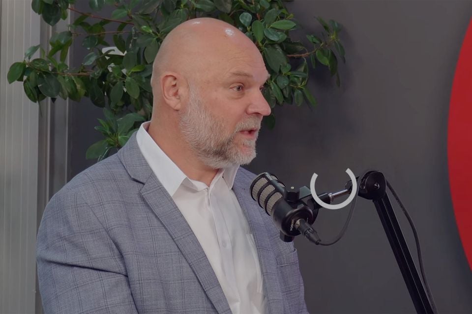 A bald man with a grey beard, wearing a grey checked blazer, speaks into a podcast microphone