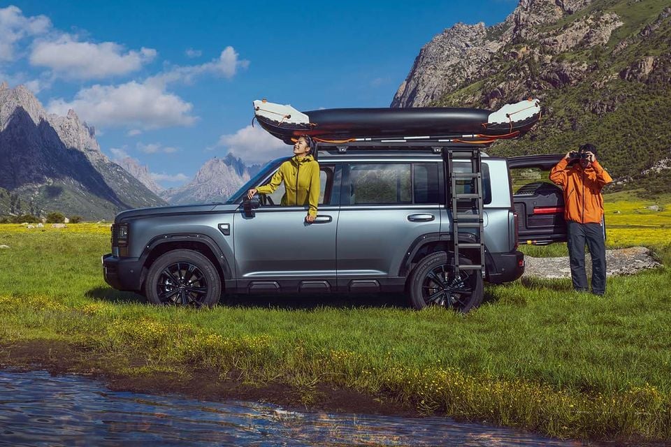 A grey Haval Raptor Menglong H7 SUV parked in an alpine meadow with a kayak strapped to the roof rack and a side ladder, with two people enjoying the outdoors in a mountainous landscape