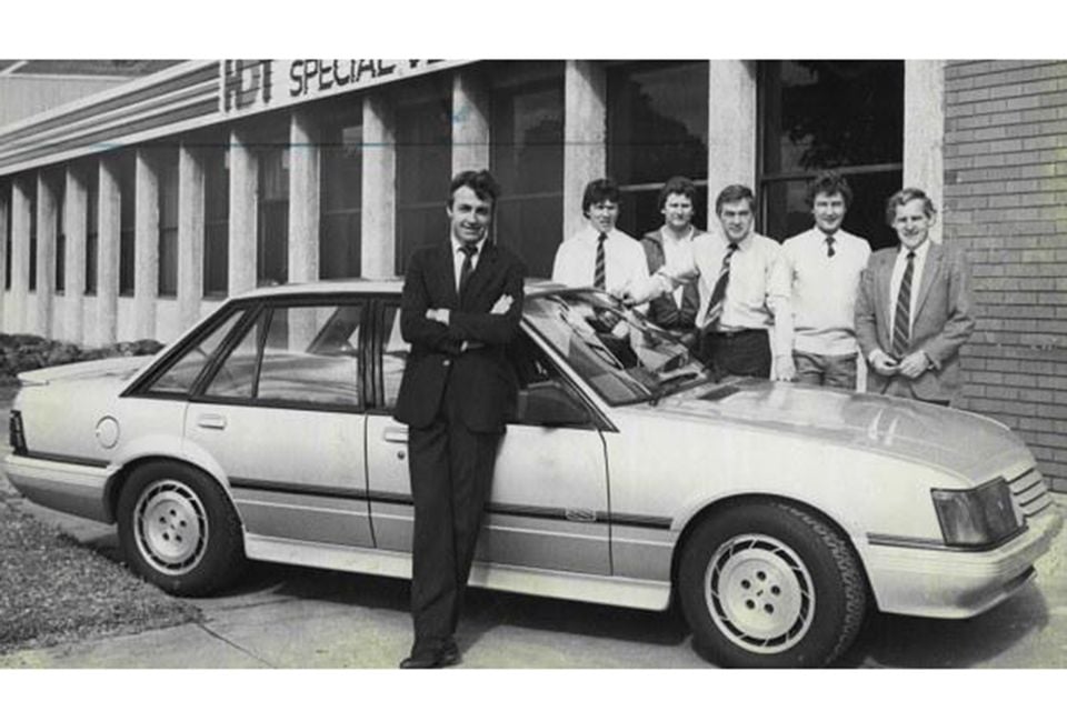 A black-and-white photo of a group of men posing with a Holden Commodore VK SS outside the HDT Special Vehicles building, believed to feature Peter Brock.