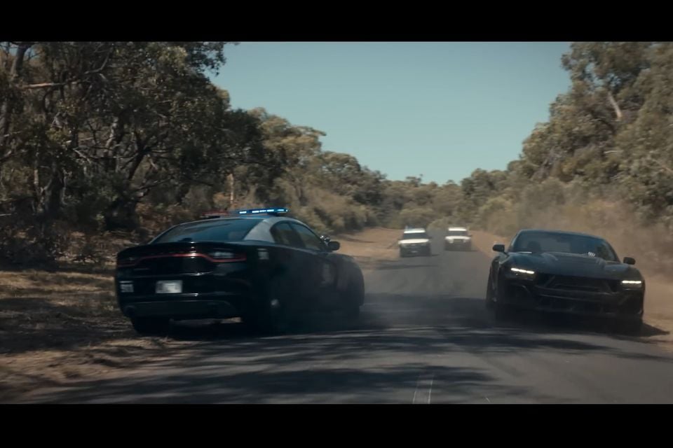 A black police car and a dark sports car racing side by side on a dusty rural road lined with Australian bush, with more vehicles visible in the distance — a cinematic film scene
