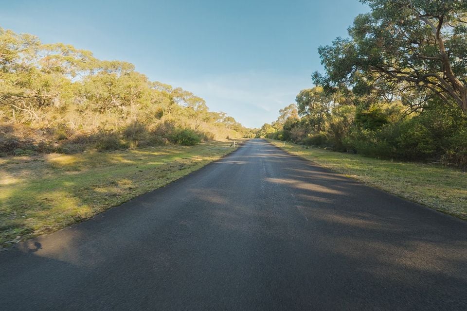 A quiet two-lane asphalt road stretching into the distance through Australian bushland under a clear blue sky
