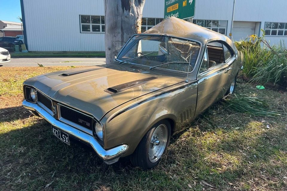 A gold Holden Monaro GTS coupe parked on grass near a tree, seen from the front-side, with a cracked windscreen suggesting crash damage