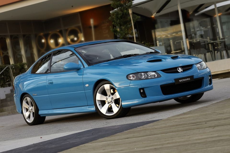 Um cupê Holden Monaro VZ azul estacionado em frente a um edifício moderno, visto de um ângulo frontal de três quartos