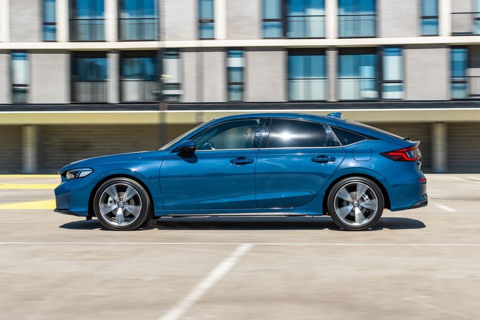 A blue Honda Civic e:HEV sedan seen from the side while driving through an urban car park area