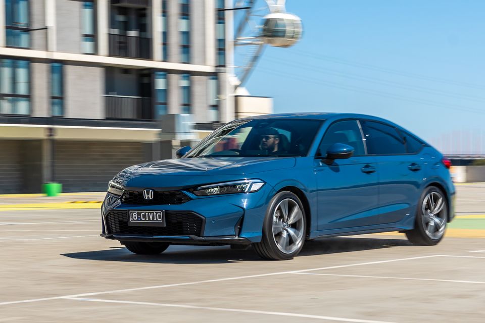 A blue Honda Civic hatchback seen from the front-side angle, driving through an open car park with urban buildings in the background