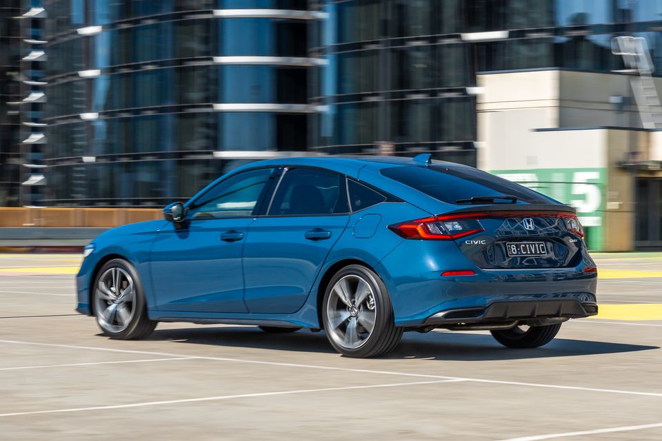A blue Honda Civic e:HEV hatchback seen from the rear three-quarter angle in an urban car park setting