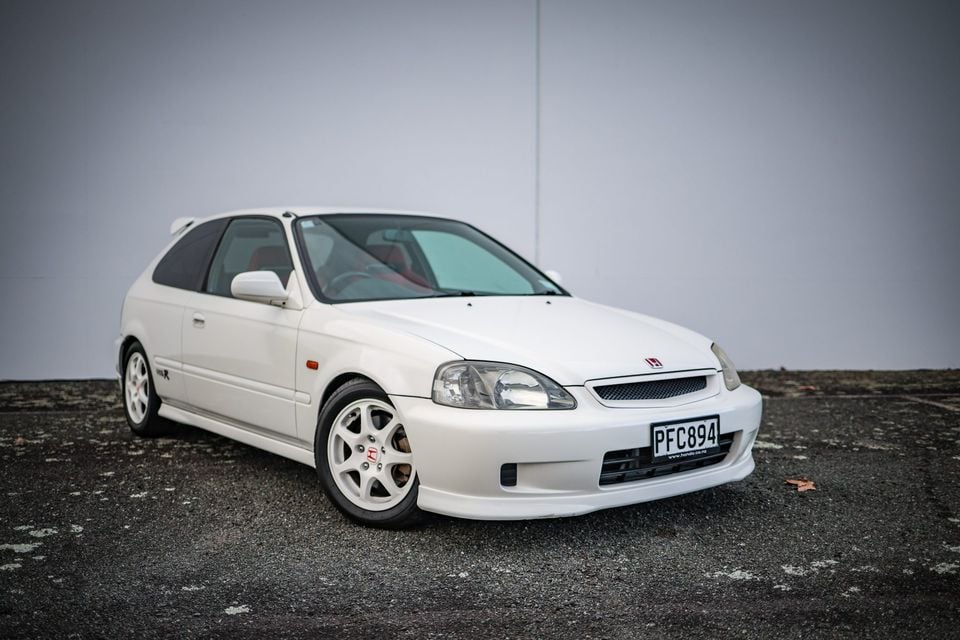 A white Honda Civic Type R hatchback seen from the front-left side, parked against a plain grey wall