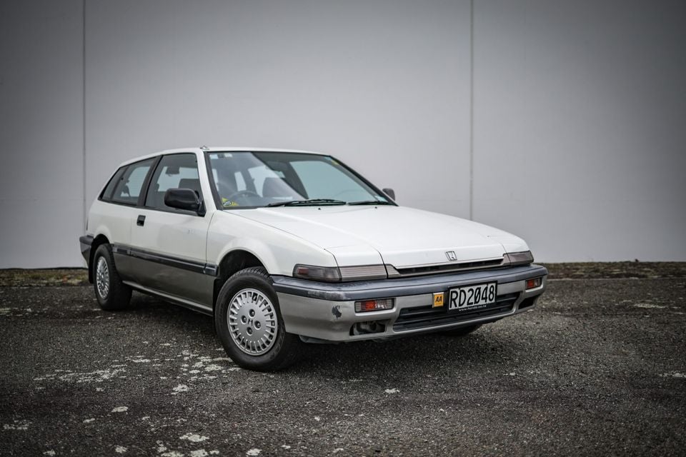 A white Honda hatchback, likely an Accord or Integra from the late 1980s, parked in front of a grey wall