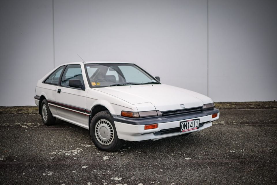 A white Honda Accord coupe parked outdoors against a plain grey wall, seen from the front-left side