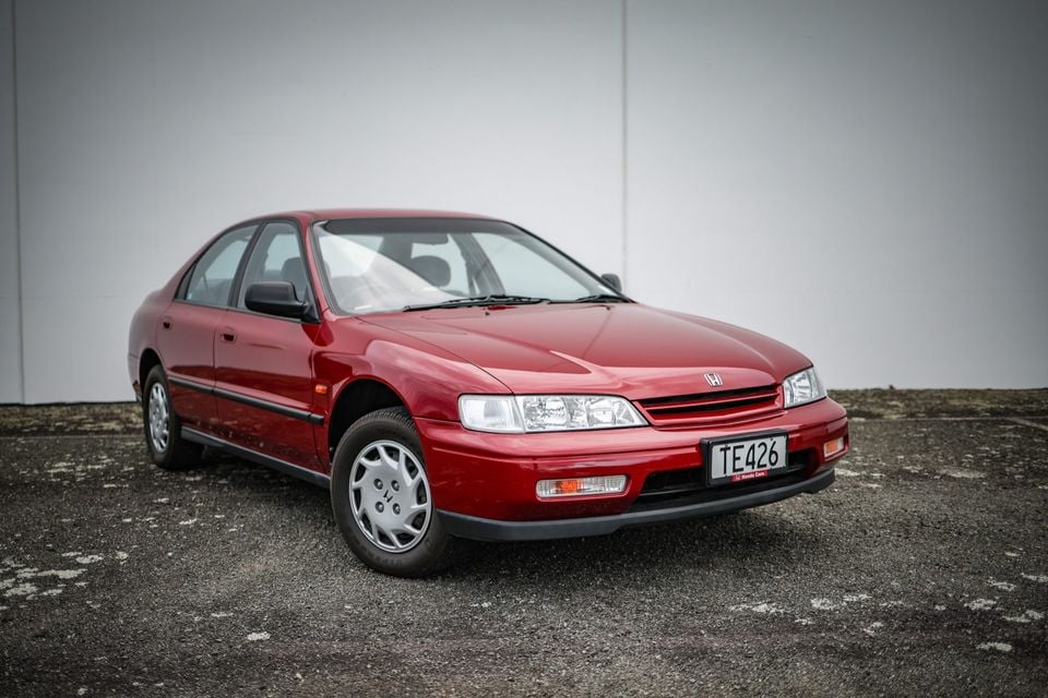A red Honda Accord sedan seen from the front-left side, parked in front of a grey wall