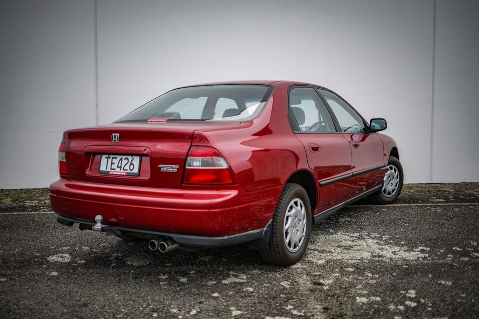 A red Honda Accord sedan seen from the rear three-quarter angle, parked in an outdoor area against a plain wall