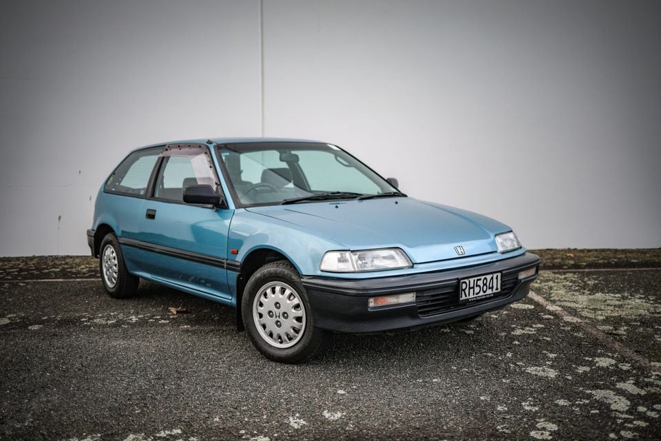 A light blue Honda Civic three-door hatchback parked on a concrete surface against a plain wall, seen from the front-left side