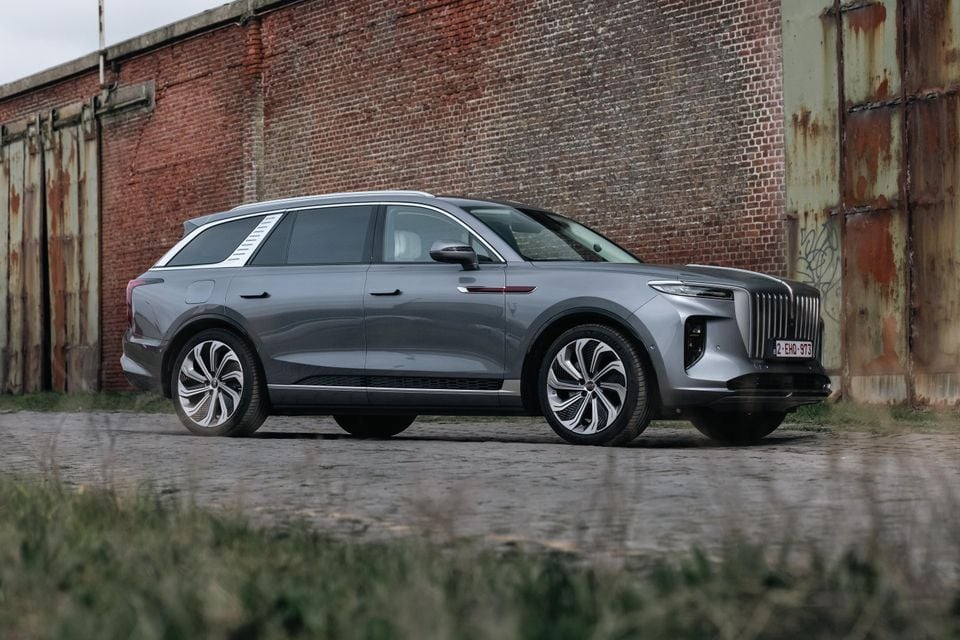 A grey Hongqi E-HS9 large SUV seen from the front-side angle, parked in front of a rustic brick wall