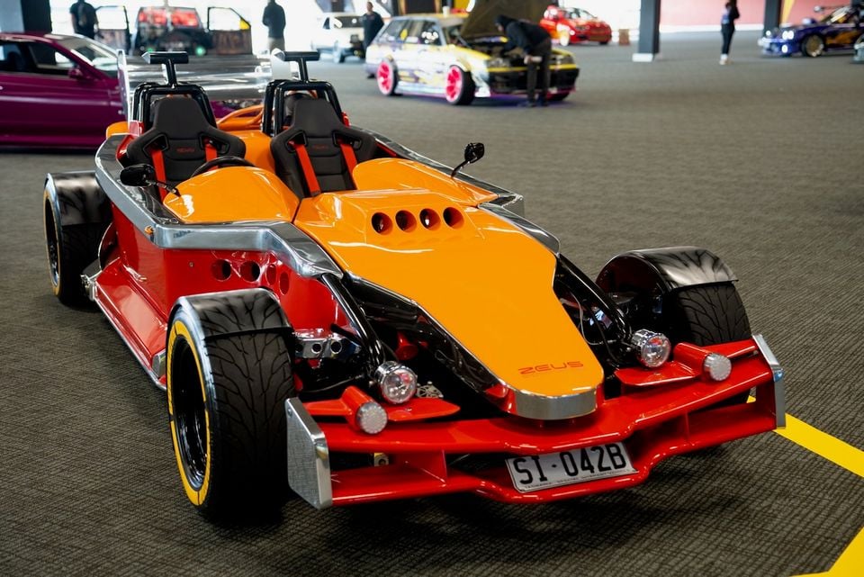 An orange and red Zeus Dominator open-top sports car on display at an indoor car show, seen from the front three-quarter angle