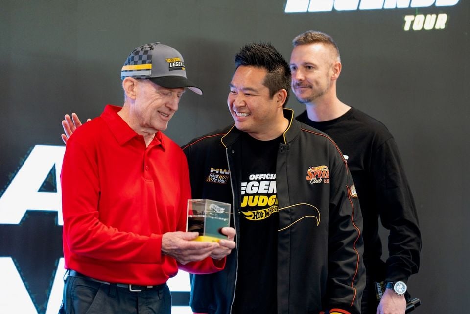 Three men pose at a Hot Wheels Legends Tour event, with one holding a trophy and another wearing an Official Legends Judge Hot Wheels jacket.