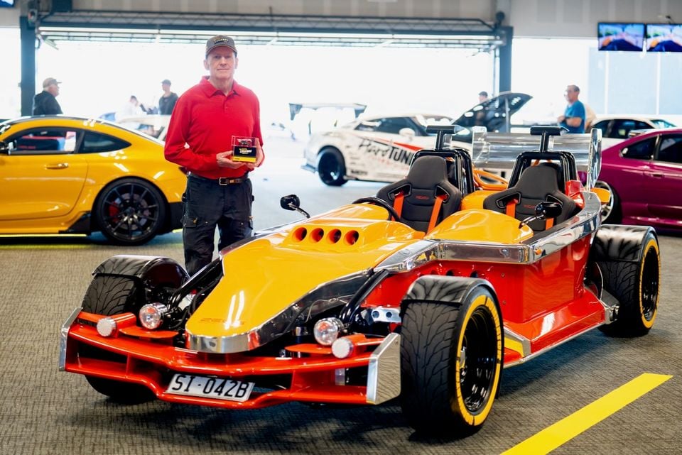 A man in a red shirt holds a trophy beside a yellow and red open-wheel kit car on display at an indoor car show, with other vehicles visible in the background