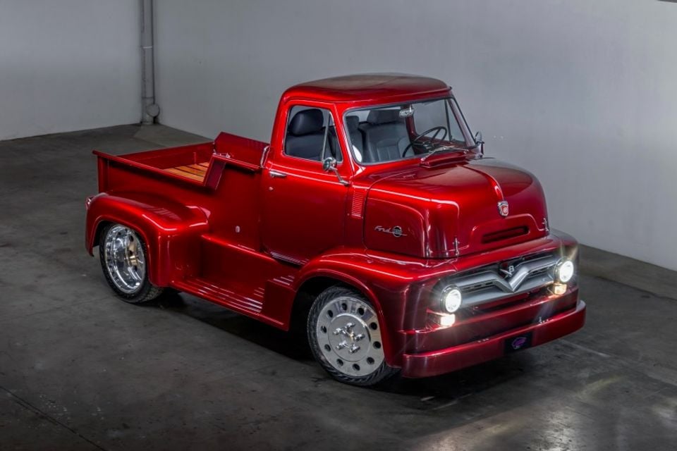 A heavily customised red vintage Ford C-series cab-over pickup truck with a candy red finish, polished chrome wheels, and lit headlights, displayed indoors