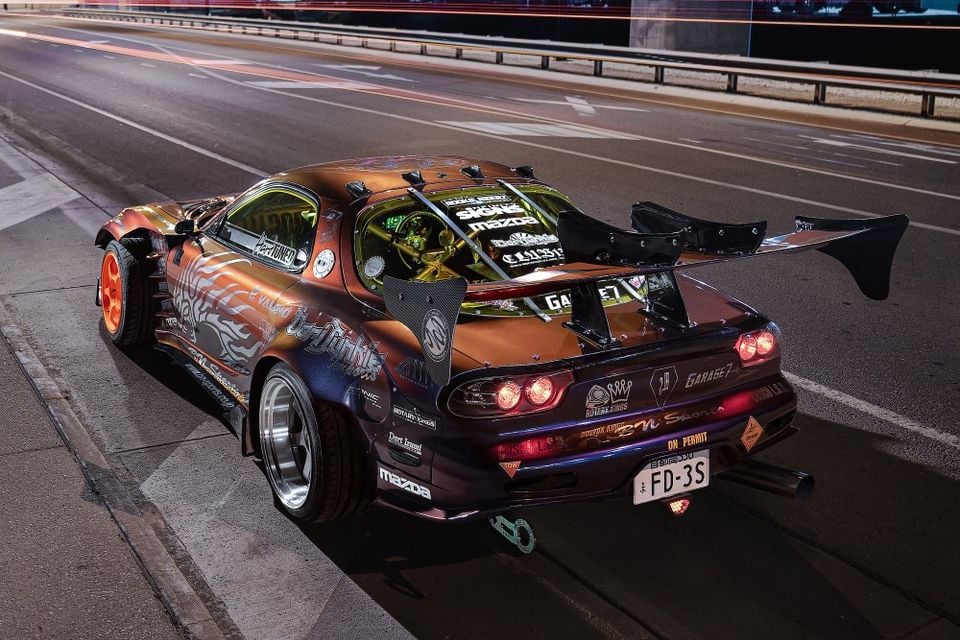A heavily modified Mazda RX-7 FD3S coupe with a wide-body kit, large rear wing, and custom bronze and purple livery, seen from the rear three-quarter angle at night on a road